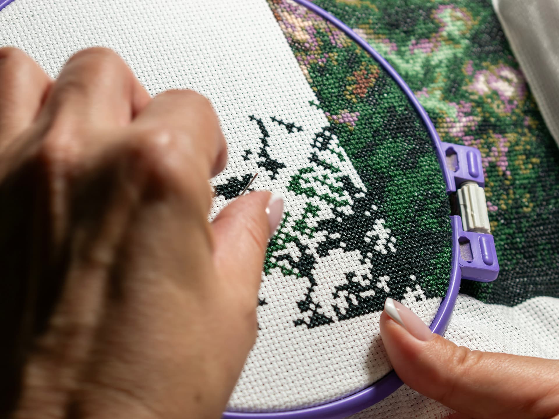 Two hands at the hoop, a needle setting a dark green cross into the Aida canvas